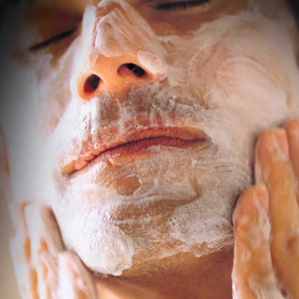 Nahaufnahme eines Mannes, der sein Gesicht mit einem schäumenden Reinigungsprodukt wäscht; geschlossene Augen, sanfte kreisende Handbewegungen und dichter, weisser Schaum auf der Haut.