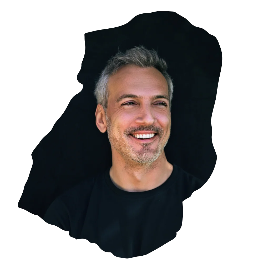 Ein lächelnder Mann mit kurzem grauem Haar und Bart blickt leicht zur Seite; er trägt ein schwarzes T-Shirt vor schwarzem Hintergrund und wirkt entspannt sowie gepflegt.